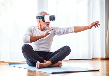 Ein älterer Mann mit grauem Haar und Bart sitzt im Schneidersitz auf einer Yogamatte in einem hellen Raum. Er trägt eine Virtual-Reality-Brille und hat die Arme ausgestreckt, als ob er eine Interaktion in der virtuellen Welt erlebt.