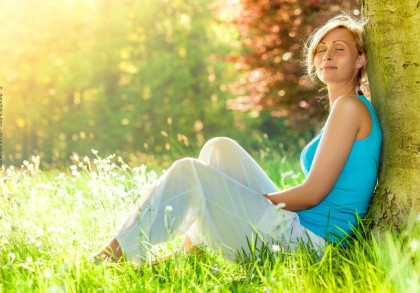 Eine Frau in einem blauen Top und weißen Hosen lehnt sich mit geschlossenen Augen an einen Baum, während sie in einer sonnendurchfluteten Wiese voller Gras und Blüten sitzt.
