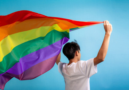Eine Person mit kurzen, dunklen Haaren steht vor einem blauen Hintergrund und hält eine große Regenbogenflagge, die im Wind flattert, über ihrem Kopf. Die Person trägt ein weißes T-Shirt und ist mit dem Rücken zur Kamera gewandt.
