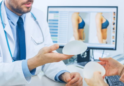 Ein Arzt im weißen Kittel zeigt einer Patientin zwei Silikonimplantate. Im Hintergrund ist ein Monitor mit Vorher-Nachher-Bildern einer Brustvergrößerung zu sehen.