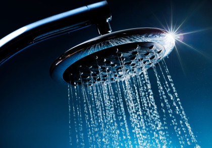 Nahaufnahme eines modernen Duschkopfes, aus dem Wasser strömt. Das Licht bricht sich an den Wassertropfen und erzeugt Reflexionen. Der Hintergrund ist dunkelblau gehalten.