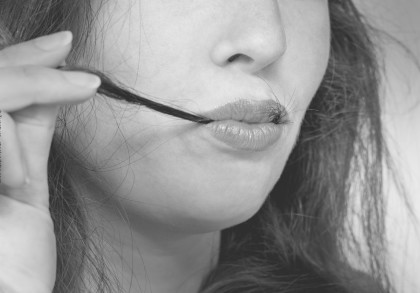 Schwarz-Weiß-Nahaufnahme einer Person, die eine Strähne ihres Haares mit den Fingern hält und es mit den Lippen berührt. Die Lippen sind leicht geschlossen und glänzend. Ein Teil des Gesichts und der Haare ist sichtbar.