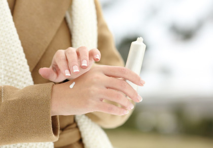 Eine Person trägt eine Creme aus einer Tube auf ihre Hand auf. Sie hat gepflegte, lackierte Fingernägel. Die Person trägt einen beigen Mantel und einen weißen Schal. Der Hintergrund ist unscharf und zeigt eine winterliche Szene.