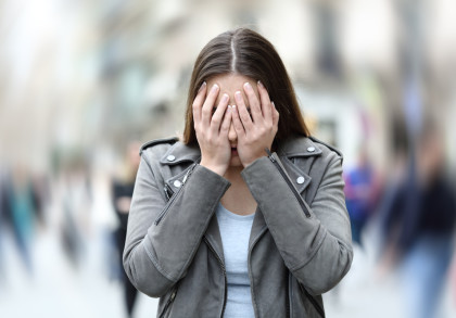 Eine Frau in einer grauen Jacke steht draußen in einer verschwommenen Umgebung und bedeckt ihr Gesicht mit ihren Händen.
