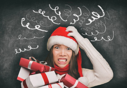Eine Frau trägt eine Weihnachtsmütze und hält mehrere Geschenke in den Händen. Sie sieht gestresst aus und hat ein verängstigtes Gesicht. Im Hintergrund ist eine Tafel mit Kritzeleien zu sehen, die Verwirrung und Stress symbolisieren.