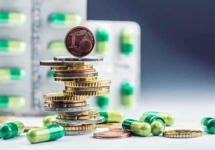 Ein Stapel Euro-Münzen mit einer Ein-Cent-Münze an der Spitze, umgeben von grünen und weißen Kapseln. Im Hintergrund sind unscharfe Blisterpackungen mit ähnlichen Kapseln zu sehen.