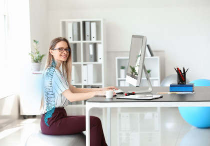 Eine lächelnde Frau sitzt auf einem Gymnastikball an einem Schreibtisch. Sie trägt eine Brille und tippt auf einem Computer. Neben ihr stehen eine Tasse, Notizbücher und Stifte. Im Hintergrund sind Regale mit Ordnern und eine Pflanze.