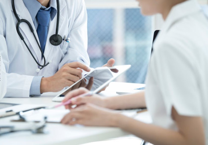Ein Arzt und ein Patient sitzen an einem Tisch in einem hell erleuchteten Raum. Der Arzt trägt einen weißen Kittel und ein Stethoskop um den Hals, während er auf ein Tablet zeigt, das der Patient hält. Auf dem Tisch: Dokumente & medizinische Instrumente.