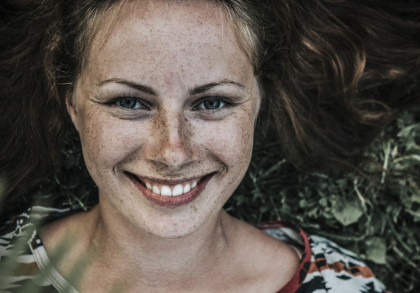 Nahaufnahme eines lächelnden Gesichts einer Frau mit hellen Augen, Sommersprossen und langen braunen Haaren. Sie liegt auf dem Rücken im Gras und trägt ein buntes Oberteil.