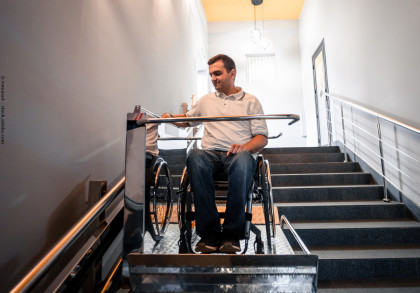 Ein Mann im Rollstuhl benutzt einen Treppenlift, um eine Treppe im Inneren eines Gebäudes hinunterzufahren. Er trägt ein weißes Polohemd und blaue Jeans. Die Treppe hat ein Metallgeländer auf beiden Seiten und die Wände sind hell gestrichen.