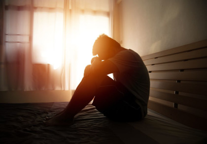 Ein Mann sitzt im Bett mit angewinkelten Beinen und stützt seinen Kopf auf seine Knie. Das Schlafzimmer ist in warmes Licht getaucht, das durch ein Fenster mit Vorhängen im Hintergrund scheint.