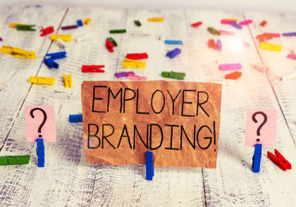 Ein Schild aus braunem Papier mit dem handgeschriebenen Text "EMPLOYER BRANDING!" steht auf einer weiß verwitterten Holzoberfläche, die mit bunten Wäscheklammern dekoriert ist.