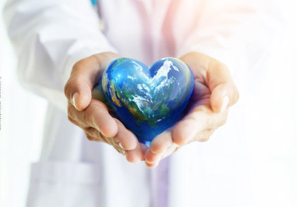 Ein Arzt in weißem Kittel hält ein herzförmiges Modell der Erde mit beiden Händen vor sich. Die Weltkugel zeigt Ozeane und Kontinente, die Landschaft ist in blauen und grünen Farben gestaltet.