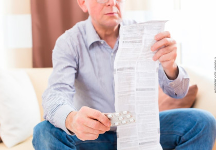 Ein älterer Mann sitzt auf einem Sofa und liest die Packungsbeilage eines Medikaments. In der rechten Hand hält er eine Blisterpackung mit Tabletten. Er trägt ein gestreiftes Hemd und eine Brille. Im Hintergrund sind eine Couch und ein Vorhang zu sehen.