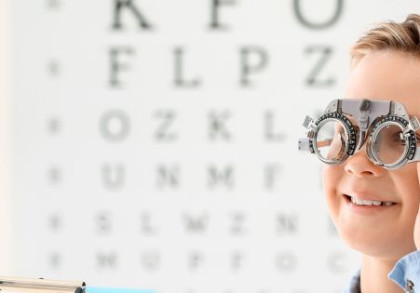 Ein Junge steht vor einer Tafel mit unterschiedlich großen Buchstaben und hat eine Brille vom Augenarzt auf.