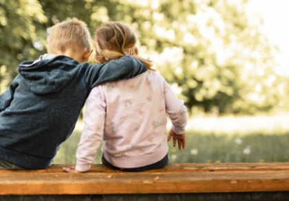 Zwei Kinder sitzen mit auf einer Bank und blicken auf eine Wiese. Der kleine Junge legt den Arm um seine Schwester.