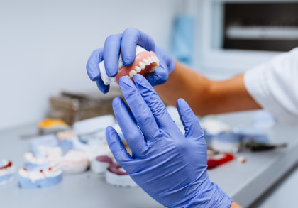 Arbeit an einer Zahnprothese. Hände in blauen Handschuhen überprüfen eine fertige Zahnprothese für den Oberkiefer. Im Hintergrund weitere Arbeitsstücke.