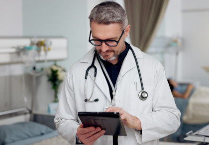 Ein Arzt steht in einem Patientenzimmer und tippt auf dem Tablet. Im Hintergrund liegt eine Patientin im Bett, eine Person in OP-Kleidung spricht mit ihr.