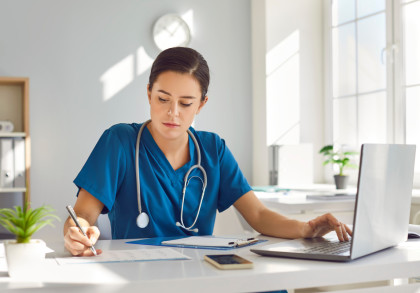 Ärztin in OP-Kleidung mit Stethoskop sitzt am Schreibtisch. Sie tippt links am Computer und schreibt mit  rechts etwas auf ein Papier.