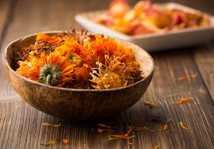 Eine hölzerne Schale mit getrockneten orangefarbenen Blütenblättern steht auf einem hölzernen Tisch. Im Hintergrund befindet sich ein unscharfes weißes Tablett mit weiteren getrockneten Blüten. Einzelne Blütenblätter sind verstreut auf dem Tisch.