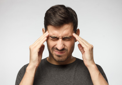 Ein braunhaariger Mann hält sich beide Hände an sein schmerzverzerrtes Gesicht, um seine Schläfen zu massieren. Er kneift dabei beide Augen fest zusammen.