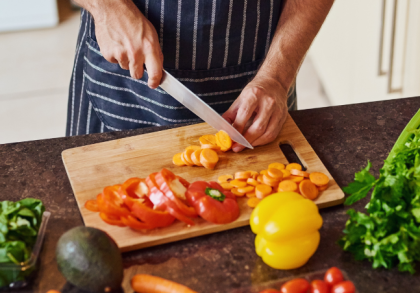 Ein Mann schneidet auf einem Küchenbrett frisches Gemüse wie Karotten und Paprika, umgeben von weiteren Zutaten wie Sellerie, Spinat, Avocado und Tomaten auf einer Küchenarbeitsplatte.
