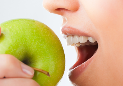 Eine Person hält einen grünen Apfel und beißt in ihn. Der Fokus liegt auf den gesunden, weißen Zähnen, die beim Kauen sichtbar sind. Das Bild zeigt die Bedeutung von Ernährung für starke Zähne.