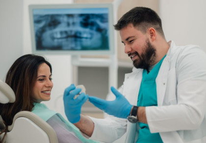 Ein Zahnarzt erklärt einer Patientin eine Behandlung anhand eines Kiefermodells. Im Hintergrund Röntgenbilder des Kiefers.