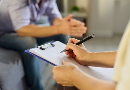 Therapeutin macht Notizen auf einem Klemmbrett während eines Gesprächs mit einem Patienten in einer Beratungssituation zur Burnout-Behandlung und Stressbewältigung in einer ruhigen Praxisumgebung.