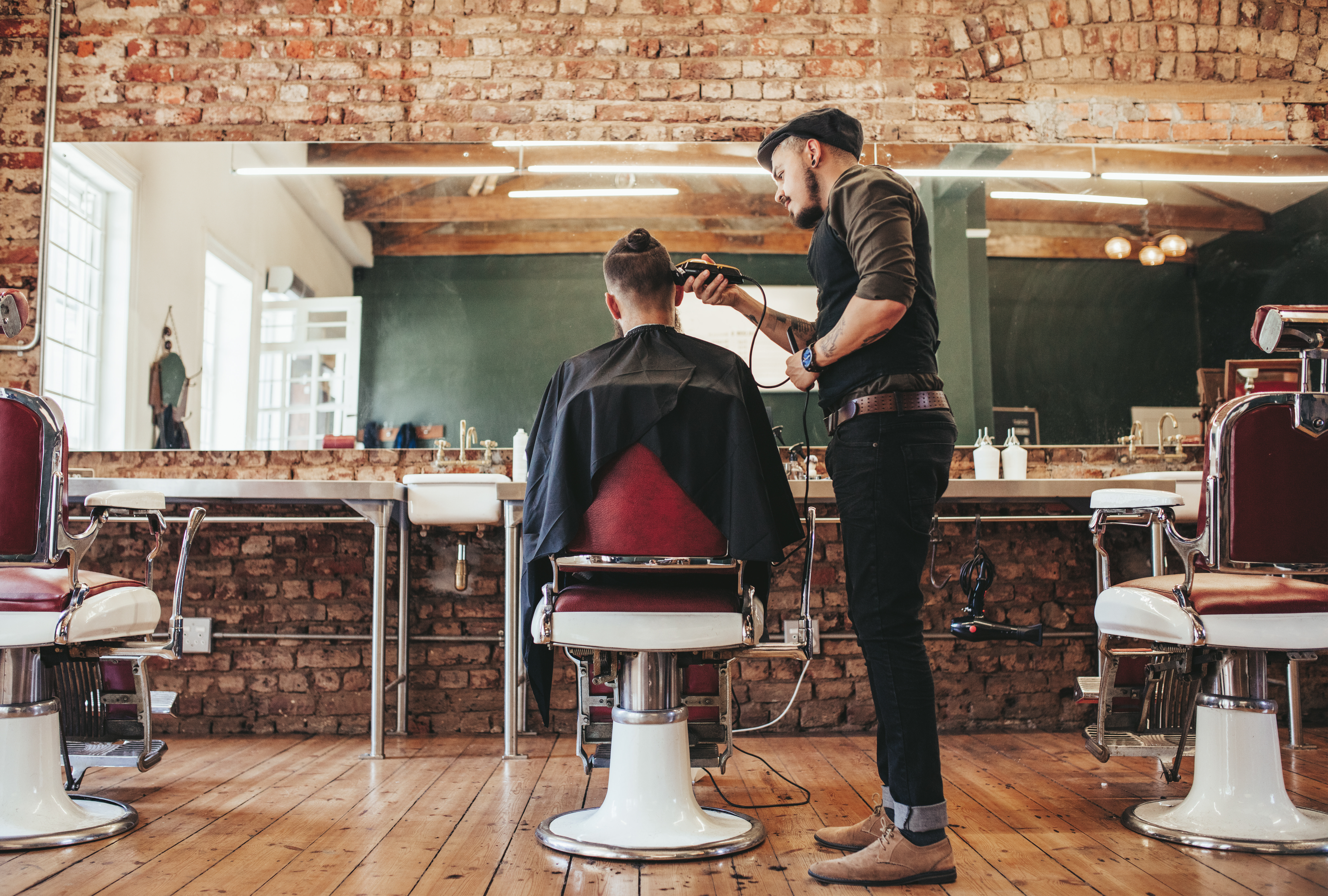 Einem Mann werden von einem anderen Mann die Haare abrasiert, während er in einem Friseursalon auf einem rotem Stuhl vor einem großen Spiegel sitzt.