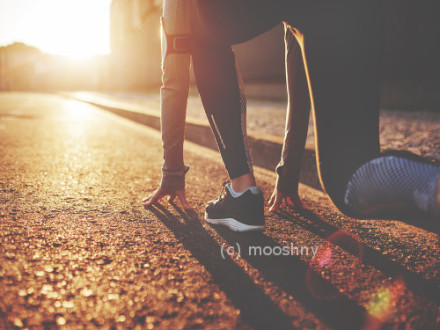 Eine Person trägt Sportkleidung und bereitet sich in einer Startposition auf einem sonnigen, asphaltierten Weg auf einen Lauf vor. Der Sonnenuntergang im Hintergrund wirft lange Schatten und taucht die Szene in ein warmes, goldenes Licht.