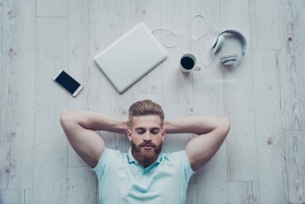 Ein Mann mit Bart und einem hellblauen T-Shirt liegt auf einem Holzboden und hat die Arme hinter dem Kopf verschränkt. Um ihn herum liegen ein geschlossenes Laptop, ein Smartphone, eine Tasse Kaffee und ein Paar Kopfhörer.