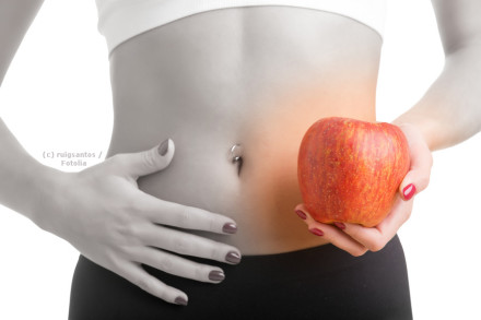 Ein farbiges Bild eines Menschen, der einen roten Apfel in der rechten Hand hält. Der Fokus liegt auf dem Bauch, der mit einem Bauchnabelpiercing verziert ist. Die linke Hand liegt auf dem Bauch.