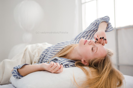 Eine Frau mit langen blonden Haaren liegt auf einem Bett und streckt sich. Sie trägt ein blau-weiß gestreiftes Oberteil und scheint entspannt zu sein. Das Zimmer ist hell und sie ruht auf einem weißen Kissen.