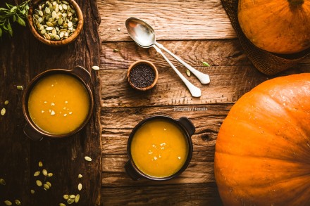 Ein rustikaler Holztisch mit zwei Schalen Kürbissuppe, umgeben von Kürbiskernen, zwei Löffeln, einer kleinen Schüssel mit schwarzen Senfkörnern und einem großen, ganzen Kürbis.