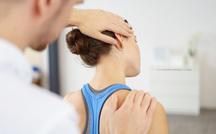 Ein Physiotherapeut behandelt den Nacken einer Frau, die ein blaues Sporttop trägt. Sie sitzt mit dem Rücken zur Kamera, während der Therapeut eine Hand an ihrem Kopf und die andere an ihrer Schulter hat.