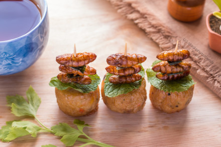 Drei Vorspeisen werden auf einem Holzbrett präsentiert. Jede Vorspeise besteht aus einem frittierten Teigbällchen, garniert mit einem frischen grünen Blatt und mehreren aufgespießten, essbaren Insekten.