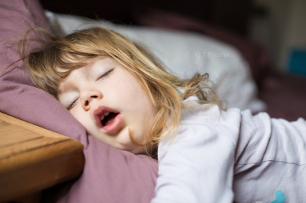 Das Bild zeigt ein schlafendes Kind mit langen, hellbraunen Haaren. Das Kind liegt auf einem Bett und trägt ein weißes Oberteil. Der Kopf des Kindes ruht auf einem lila Kissen und der Mund ist leicht geöffnet.