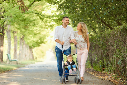 Ein junges Paar spaziert mit ihrem Kleinkind in einem Kinderwagen einen von Bäumen gesäumten Weg entlang. Der Mann trägt ein weißes Hemd und blaue Jeans, während die Frau ein blumiges T-Shirt und rosa Hosen trägt.