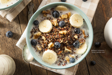 Eine Schüssel Quinoa-Frühstücksbrei mit geschnittenen Bananen, Blaubeeren und Nüssen, serviert auf einem Holztisch. Daneben liegt ein in eine Serviette gehüllter Löffel.