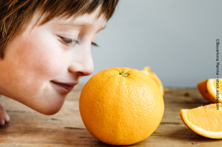 Ein Kind riecht an einer Orange, die auf einem Holztisch liegt. Mehrere Orangenscheiben sind im Hintergrund zu sehen.