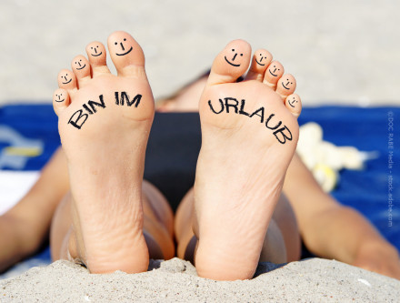 Zu sehen sind die Fußsohlen einer Person am Strand. Auf den Zehen sind lächelnde Gesichter gemalt. Auf der linken Fußsohle steht „BIN IM“ und auf der rechten „URLAUB“, zusammen „BIN IM URLAUB“. Die Person liegt entspannt auf einem blauen Handtuch.