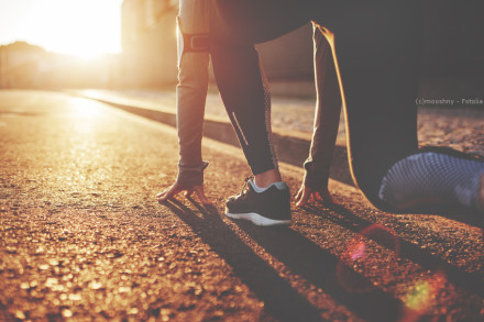 Eine Person in Sportkleidung befindet sich in der Startposition für einen Lauf auf einer Straße bei Sonnenaufgang oder Sonnenuntergang. Sie trägt schwarze Laufschuhe und die Szene ist in warmes, goldenes Licht getaucht.