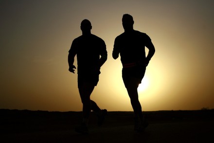 Zwei Personen joggen bei Sonnenuntergang. Ihre Silhouetten sind vor der untergehenden Sonne sichtbar. Der Himmel ist düster mit einem Hauch von Orange und Gelb.