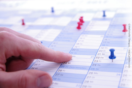 Nahaufnahme eines Fingers, der auf den 24. Mai (Mittwoch) in einem Monatskalender zeigt. Der Kalender enthält mehrere bunte Stecknadeln (rot und blau), die verschiedene Daten markieren. Der Hintergrund ist unscharf.