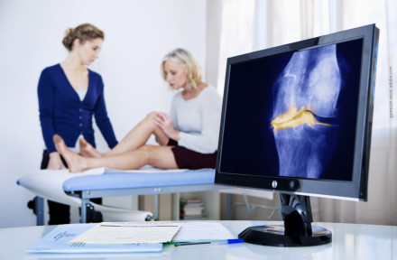 Ein Arzt untersucht das Knie einer Patientin, die auf einem Untersuchungstisch sitzt. Im Vordergrund auf einem Schreibtisch steht ein Monitor, auf dem eine Röntgenaufnahme eines Knies zu sehen ist. Daneben liegen einige Dokumente und ein Kugelschreiber.