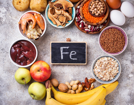 Eine Auswahl verschiedener eisenreicher Lebensmittel, darunter Kartoffeln, Garnelen, getrocknete Pilze, Hülsenfrüchte, Eier, Leber, Äpfel, Bananen, Nüsse & Haferflocken. In der Mitte steht eine kleine Tafel mit der chemischen Bezeichnung für Eisen, "Fe".
