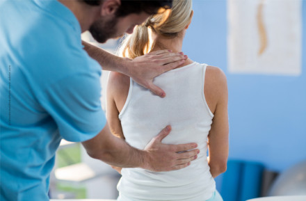 Ein Physiotherapeut behandelt den oberen Rücken einer Frau in einem Behandlungsraum. Die Frau trägt ein weißes Tanktop und sitzt mit dem Rücken zur Kamera, während der Therapeut in einem blauen Hemd seine Hände auf ihren Rücken legt.