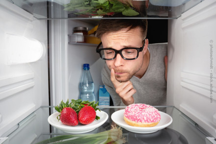 Ein Mann mit Brille schaut nachdenklich in einen Kühlschrank. Im Vordergrund befinden sich auf einem Regal zwei Teller, einer mit frischen Erdbeeren & einer mit einem rosa glasierten Donut. Im Hintergrund stehen eine Wasserflasche und andere Lebensmittel.