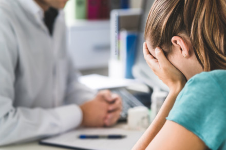 Eine Frau sitzt mit dem Gesicht in den Händen und scheint verzweifelt oder traurig zu sein. Vor ihr sitzt eine Person, wahrscheinlich ein Arzt oder Berater, der seine Hände gefaltet vor sich auf dem Tisch liegen hat.